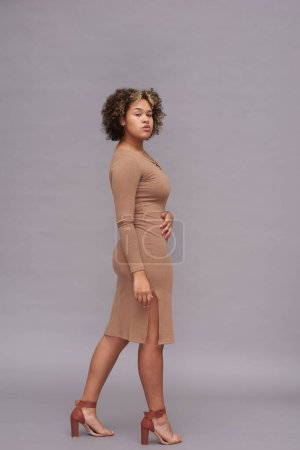 Téléchargez les photos : Jeune femme afro-américaine en robe décontractée de couleur beige vous regardant tout en posant devant la caméra contre le mur gris en studio - en image libre de droit