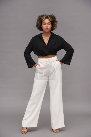 Téléchargez les photos : Jeune femme multi-ethnique confiante en haut recadré noir et pantalon blanc gardant les mains dans les poches tout en posant devant la caméra - en image libre de droit