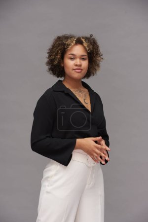 Foto de Mujer joven multiétnica en ropa casual inteligente de pie frente a la cámara contra el fondo gris durante la sesión de fotos - Imagen libre de derechos