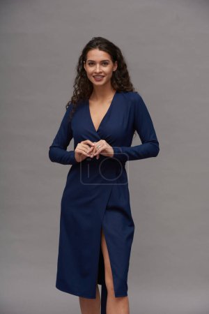 Photo for Gorgeous brunette woman with long hair wearing dark blue elegant dress looking at camera with smile while standing against grey wall - Royalty Free Image