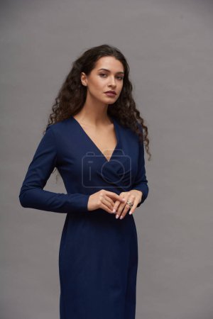 Téléchargez les photos : Jeune femme aux cheveux longs foncés portant une robe bleue intelligente lors d'une séance photo en studio tout en se tenant debout sur fond gris - en image libre de droit