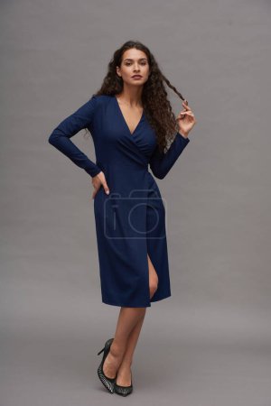 Téléchargez les photos : Jeune femme séduisante en robe bleue et chaussures à talons hauts posant pour appareil photo contre un mur gris lors d'une séance photo en studio - en image libre de droit