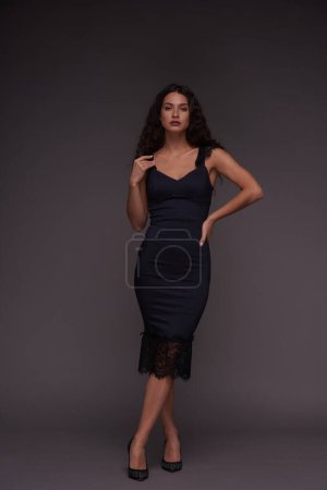 Photo for Young lady in black silk dress and high heeled shoes posing during photo session while standing over grey wall and looking at camera - Royalty Free Image