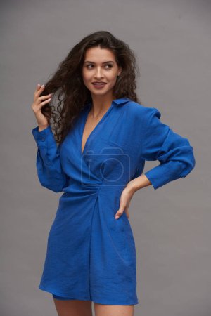 Photo for Happy young brunette fashion model in smart blue dress touching her dark long wavy hair and looking aside during photo session - Royalty Free Image