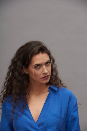 Téléchargez les photos : Jeune femme brune avec de longs cheveux foncés ondulés debout devant la caméra tout en posant pendant la séance photo sur fond gris - en image libre de droit
