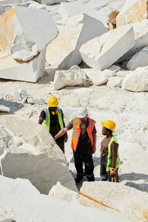 Foto de Grupo intercultural de control de calidad en ropa de trabajo de pie frente a una enorme roca de mármol blanco mientras uno de ellos lo toca - Imagen libre de derechos