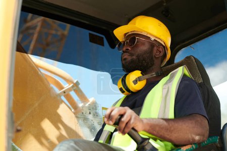 Foto de Joven conductor masculino negro de la máquina de construcción en casco de seguridad y uniforme sentado por buey y mirando a través de la ventana delantera - Imagen libre de derechos