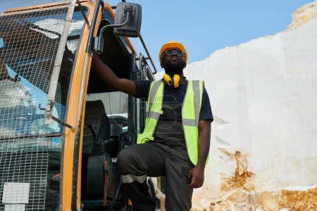 Foto de Joven hombre afroamericano en ropa de trabajo y casco de pie en la excavadora u otra máquina de construcción durante el trabajo - Imagen libre de derechos