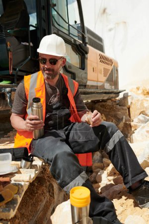 Foto de Ingeniero masculino de mediana edad en ropa de trabajo y hardhat que tiene sándwich y té caliente para el almuerzo mientras está sentado en la oruga de la excavadora - Imagen libre de derechos