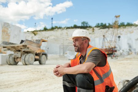 Photo for Tired worker of marble quarry sitting in front of camera by workplace against dump truck and working bulldozer with crane - Royalty Free Image
