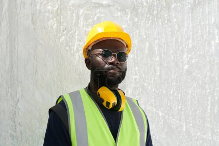 Téléchargez les photos : Jeune ingénieur afro-américain sérieux en casque de sécurité, gilet réfléchissant et lunettes regardant caméra contre mur de marbre blanc - en image libre de droit