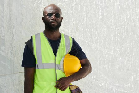 Photo for Serious black man in reflective vest and eyeglasses holding hardhat by his waist while standing in front of camera against white wall - Royalty Free Image