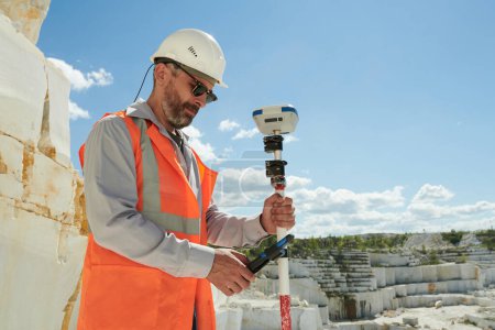 Confident engineer or surveyor with geodesic equipment standing on territory of marble quarry and adjusting theodolite