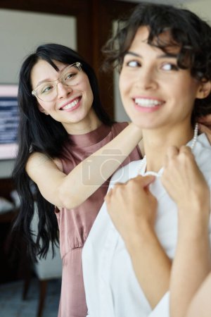 Téléchargez les photos : Joyeux jeune femme brune aidant sa sœur en peignoir de soie blanche mettre collier de perles sur le cou pendant la préparation du mariage - en image libre de droit