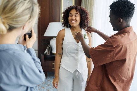 Téléchargez les photos : Joyeux jeune mariée afro-américaine en costume élégant blanc posant devant demoiselle d'honneur avec smartphone le matin du mariage - en image libre de droit