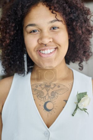 Foto de Joven mujer negra sonriente con tatuaje en el pecho en chaleco blanco con flor de ojal de pie delante de la cámara en la boda - Imagen libre de derechos
