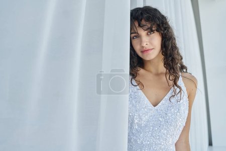 Téléchargez les photos : Jeune femme magnifique en belle et élégante robe de mariée blanche avec des paillettes debout entre les rideaux de mousseline de soie devant la caméra - en image libre de droit