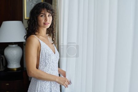 Téléchargez les photos : Jeune femme brune magnifique en robe de mariée blanche regardant la caméra tout en restant près de la fenêtre avec des rideaux en mousseline de soie - en image libre de droit