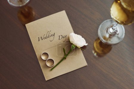 Foto de Vista superior de la composición de la boda en la mesa que consta de invitación, rosa blanca fresca y dos anillos rodeados de flautas de champán - Imagen libre de derechos