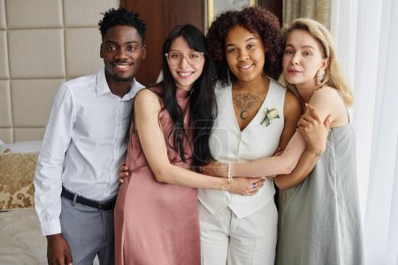 Téléchargez les photos : Heureuses jeunes femmes interculturelles et gars debout par la mariée afro-américaine en costume blanc le jour du mariage et en regardant la caméra - en image libre de droit