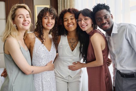 Téléchargez les photos : Groupe interculturel d'invités de mariage élégants heureux entourant les jeunes mariés affectueux en tenue blanche tout en appréciant la fête à la maison - en image libre de droit