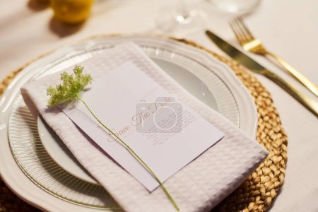 Photo for Close-up of white porcelain plate with folded cotton napkin and small green decorative plant over card with text on served table - Royalty Free Image