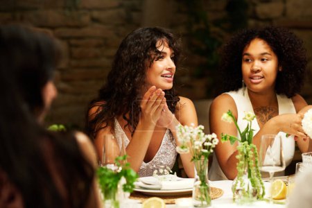 Foto de Joven pareja lésbica intercultural vestida de blanco sentada junto a la mesa festiva servida durante la fiesta de bodas y comunicándose con sus invitados - Imagen libre de derechos