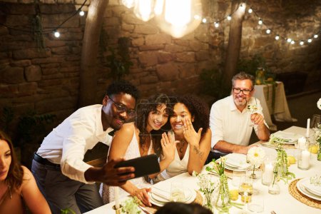 Foto de Dos novias lesbianas jactándose con sus anillos de bodas por mesa festiva servida mientras miran la pantalla del teléfono inteligente sostenida por el joven hombre negro - Imagen libre de derechos