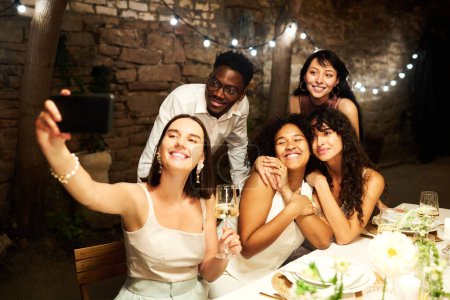 Téléchargez les photos : Groupe de jeunes amis interculturels heureux faisant selfie par table de fête servi tout en appréciant la fête de mariage dans un restaurant luxueux - en image libre de droit