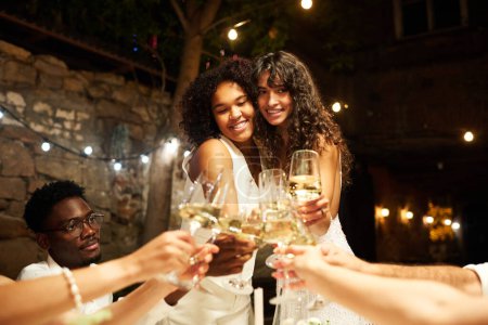 Foto de Feliz joven novias cariñosas en traje de boda tintineo con flautas de champán con sus amigos e invitados en la fiesta en el restaurante - Imagen libre de derechos