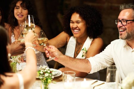 Foto de Feliz joven afroamericana novia en traje de boda brindis con amigos por la mesa festiva servida mientras disfruta de la fiesta - Imagen libre de derechos