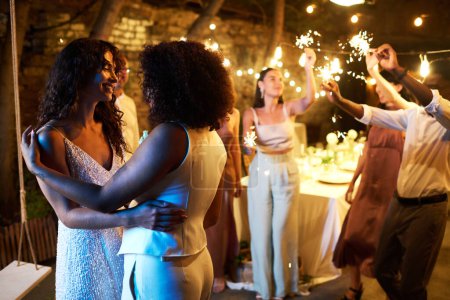 Foto de Dos jóvenes mujeres interculturales amorosas en traje de novia blanco bailando delante de la cámara contra sus amigos con bengalas divirtiéndose - Imagen libre de derechos