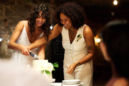 Foto de Dos jóvenes novias alegres con elegante atuendo de boda cortando un gran pastel mientras están de pie junto a la mesa festiva frente a los invitados disfrutando de la fiesta - Imagen libre de derechos