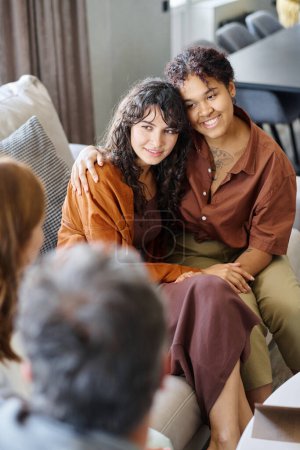 Téléchargez les photos : Joyeux jeune couple lesbien en tenue décontractée assis les uns à côté des autres sur le canapé devant les parents de l'un d'eux pendant la conversation - en image libre de droit