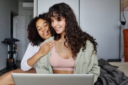 Photo for Young affectionate African American woman embracing her girlfriend sitting on double bed in front of laptop and networking - Royalty Free Image