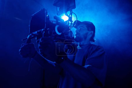 Foto de Joven en ropa de casualwear sosteniendo la cámara de vídeo mientras filma comercial en estudio iluminado con luz azul y lleno de humo - Imagen libre de derechos