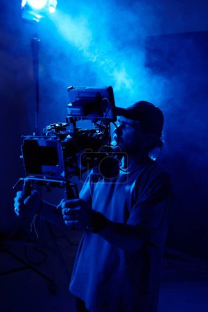 Foto de Serio joven camarógrafo con steadicam de pie contra el centro de atención en el estudio iluminado por la luz azul y lleno de humo durante la filmación de vídeo - Imagen libre de derechos