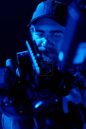 Foto de Parte de la cara del camarógrafo barbudo sosteniendo videocamera durante la filmación de un nuevo comercial en un estudio oscuro iluminado por luz azul - Imagen libre de derechos