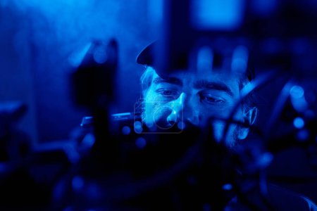 Téléchargez les photos : Partie supérieure du visage du vidéaste tenant le steadicam pendant le tournage commercial dans un studio sombre rempli de fumée et éclairé par la lumière bleue - en image libre de droit