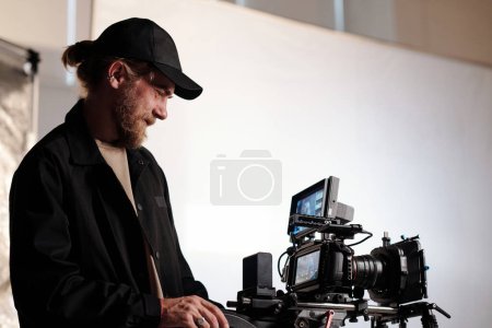 Photo for Side view portrait of bearded cameraman watching at screen of steadicam while adjusting focus and other settings before shooting commercial - Royalty Free Image