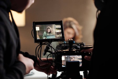 Foto de Primer plano de las pantallas steadicam con el modelo femenino usando el ordenador portátil por la mesa durante comercial que es filmado por el camarógrafo y su asistente - Imagen libre de derechos
