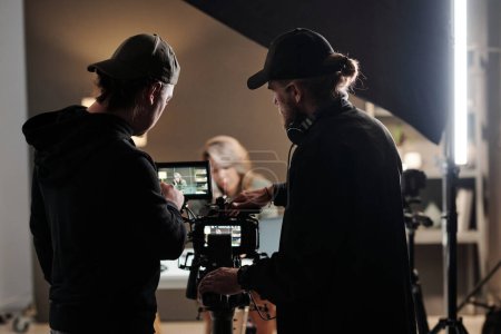 Vue arrière de deux jeunes hommes debout devant la caméra et utilisant steadicam lors du tournage d'une nouvelle publicité avec un modèle féminin