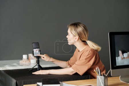 Téléchargez les photos : Jeune femme blonde regardant à travers les photos à l'écran du smartphone après la prise de vue tout en étant assis par le lieu de travail dans le studio de production - en image libre de droit