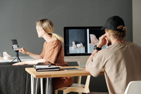 Foto de Joven fotógrafo sentado frente al monitor de la computadora y asistente femenina mirando a través de nuevas fotos en el teléfono inteligente - Imagen libre de derechos