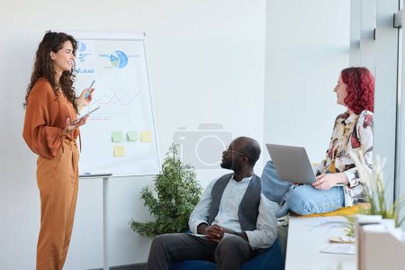 Téléchargez les photos : Jeune femme d'affaires confiante debout au tableau blanc avec des diagrammes financiers et des graphiques tout en expliquant l'un d'eux à ses collègues - en image libre de droit
