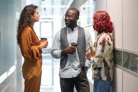 Photo for Three young successful office managers in casualwear having chat while standing between two walls in corridor of modern building - Royalty Free Image