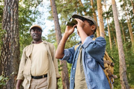 Photo for Cute youthful boy looking far away through binoculars while hiking in pine tree forest with his grandfather on summer weekend - Royalty Free Image