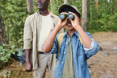 Foto de Lindo niño en ropa casual y gorra de béisbol mirando a través de los prismáticos mientras está de pie en el bosque de pinos en el día de verano contra su abuelo - Imagen libre de derechos