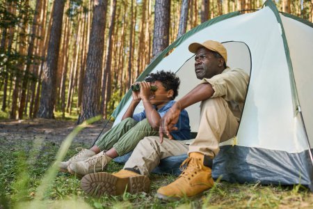 Téléchargez les photos : Mignon garçon afro-américain regardant à travers des jumelles tout en étant assis dans la tente à côté de son grand-père en tenue décontractée pendant la randonnée dans la forêt - en image libre de droit