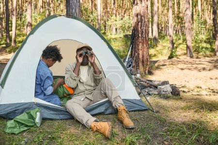 Photo for Retired African American man in casualwear looking through binoculars while sitting in tent next to his grandson unpacking rucksack - Royalty Free Image
